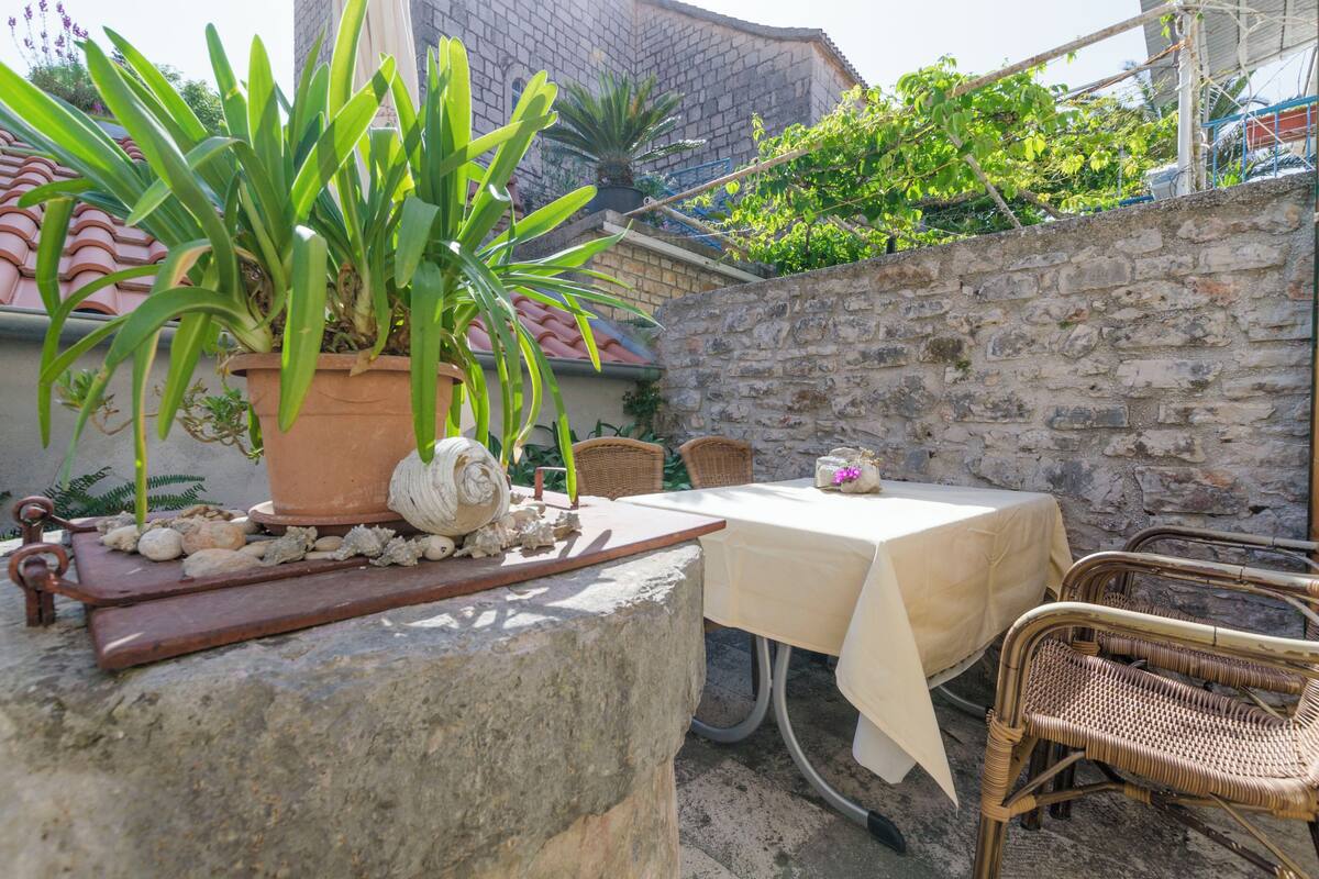 Un mur de pierre, une table recouverte d’une nappe blanche, des chaises en osier et une plante en pot.