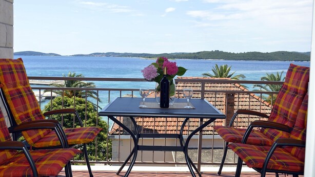 Ein Balkon mit Tisch, zwei Stühlen, einer Blumenvase und Blick auf das Meer und die Hügel.