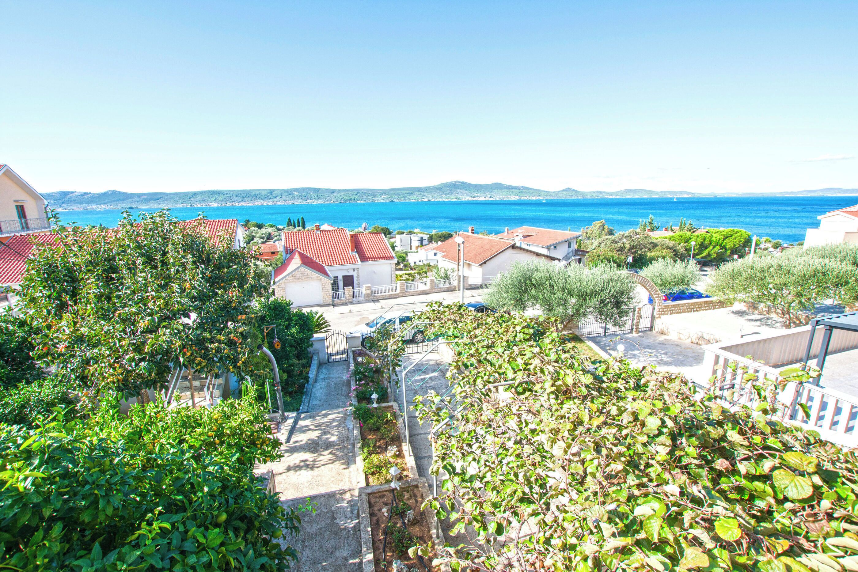 A coastal residential area with houses, a clear blue sea, and a hilly landscape in the background.