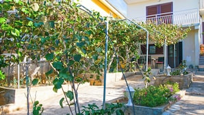 A courtyard with a climbing vine structure, a small garden area with flowers, and a building with a balcony. - Apartment Luce (Sveti Filip i Jakov)