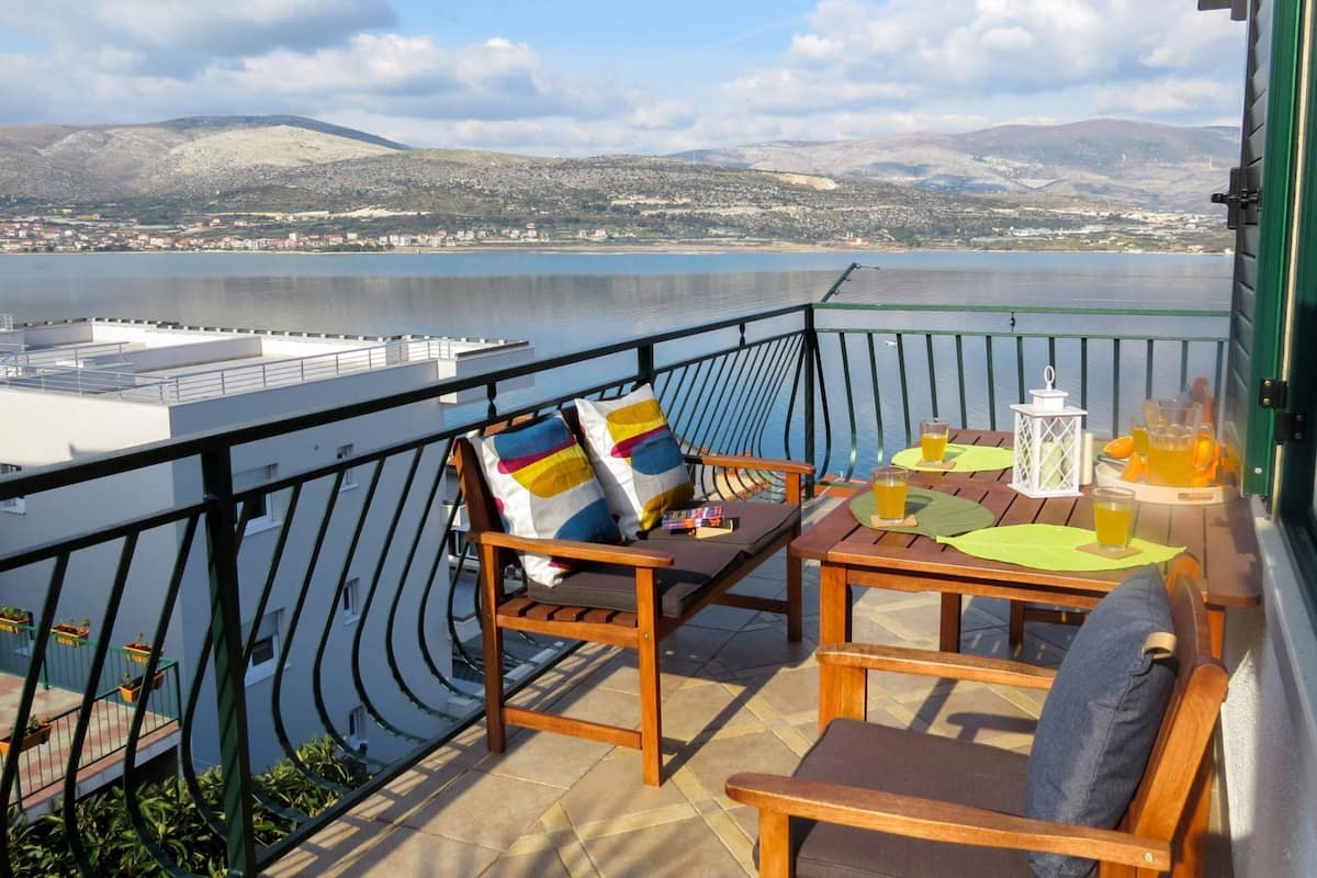 A balcony with a view of a body of water and distant hills, furnished with wooden chairs and a table with drinks.