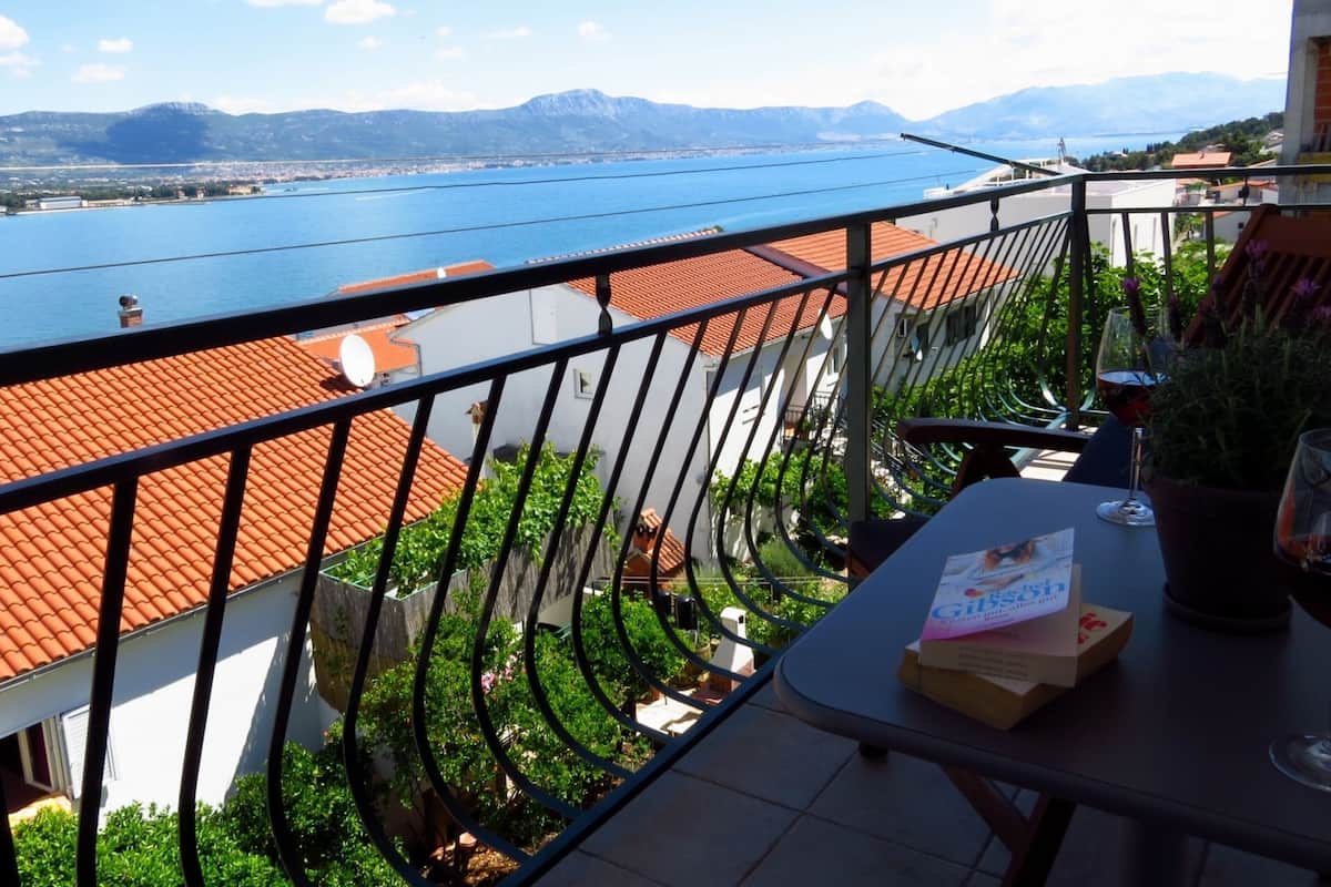 A balcony with a view of a lake and mountains, a table with a glass of wine and a book, and a potted plant.