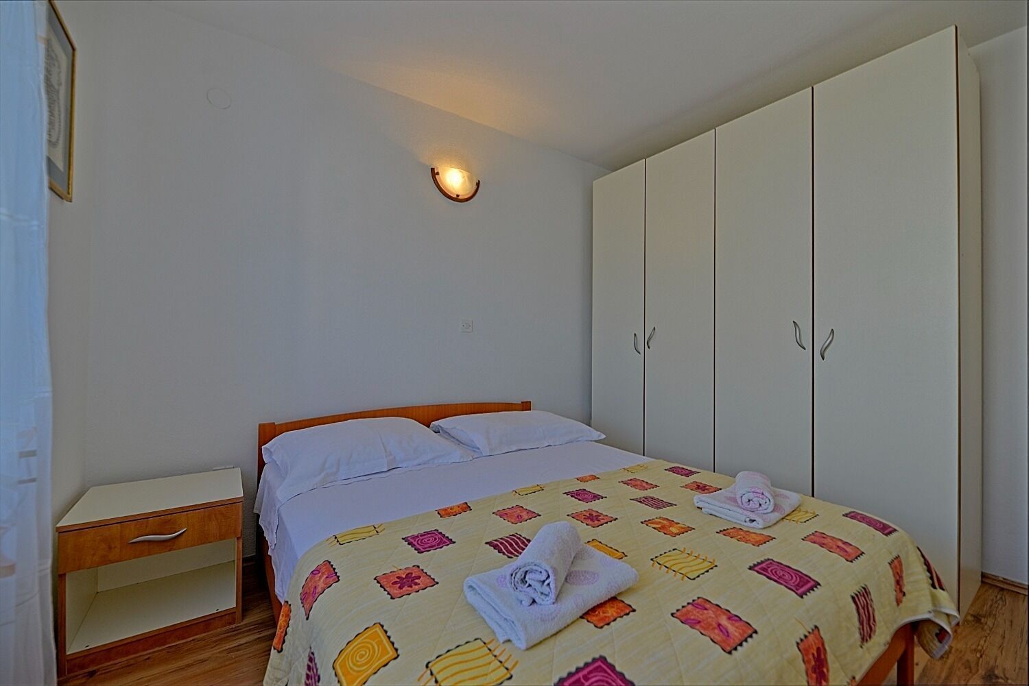 A bedroom with a bed, a nightstand, a wardrobe, and a wall-mounted light.