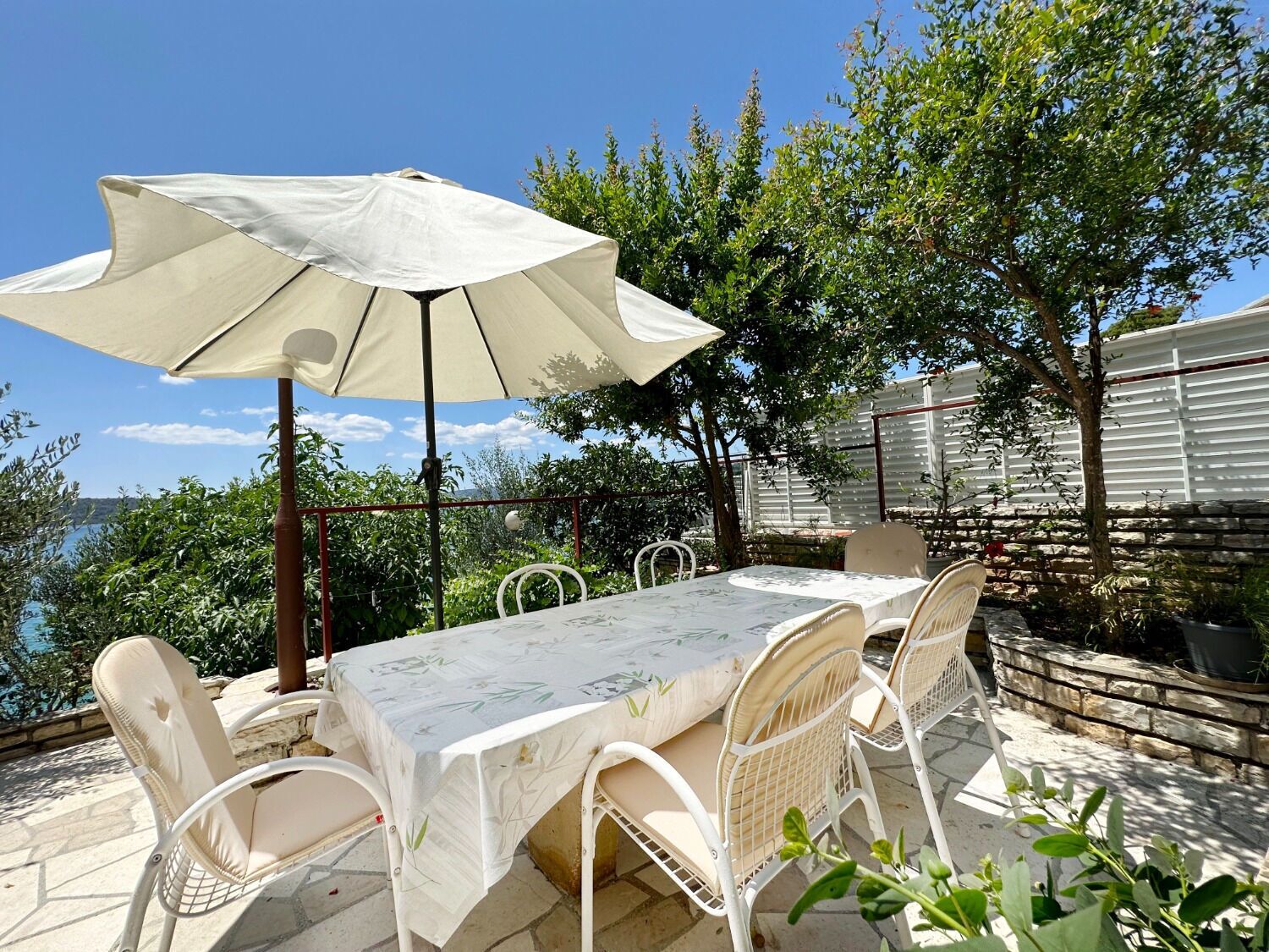 Eine Terrasse im Freien mit Tisch, Stühlen und einem großen Sonnenschirm, mit Blick auf das Meer.