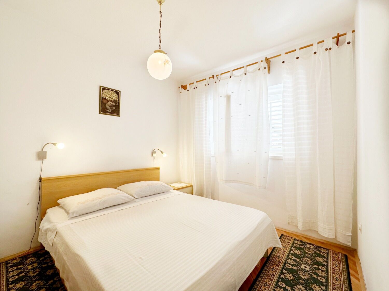 Uma cama bem arrumada com cabeceira de madeira, duas luminárias de apoio, um lustre e uma janela com cortinas leves.