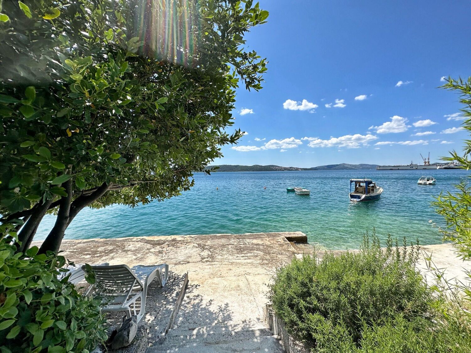 A coastal view with a clear sky, boats on the water, and a sandy area with chairs and a table.