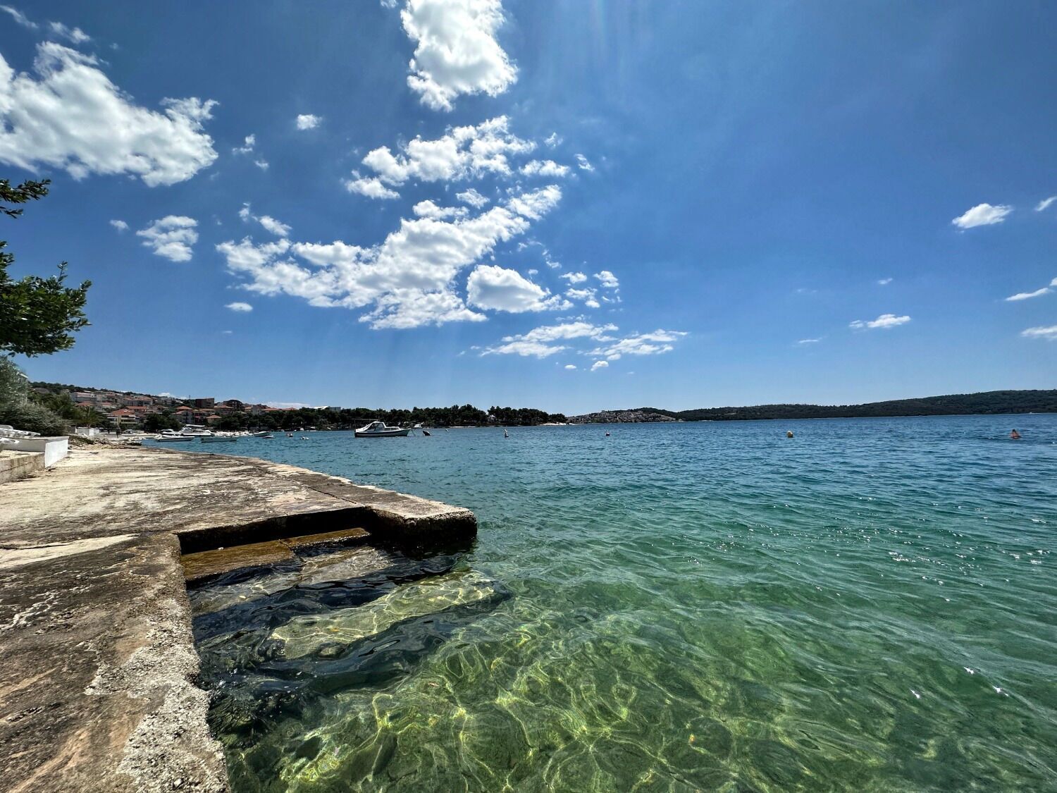 Klares Wasser mit einer Stegpromenade, ruhiger Wasserfläche und einer fernen Küste mit Gebäuden und Booten.