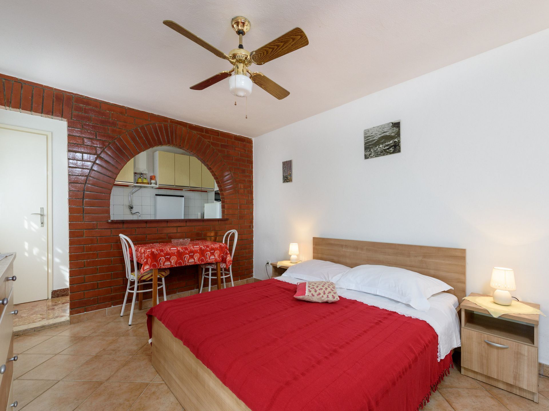 A bedroom with a bed, a nightstand, a table with chairs, a ceiling fan, and a brick wall.