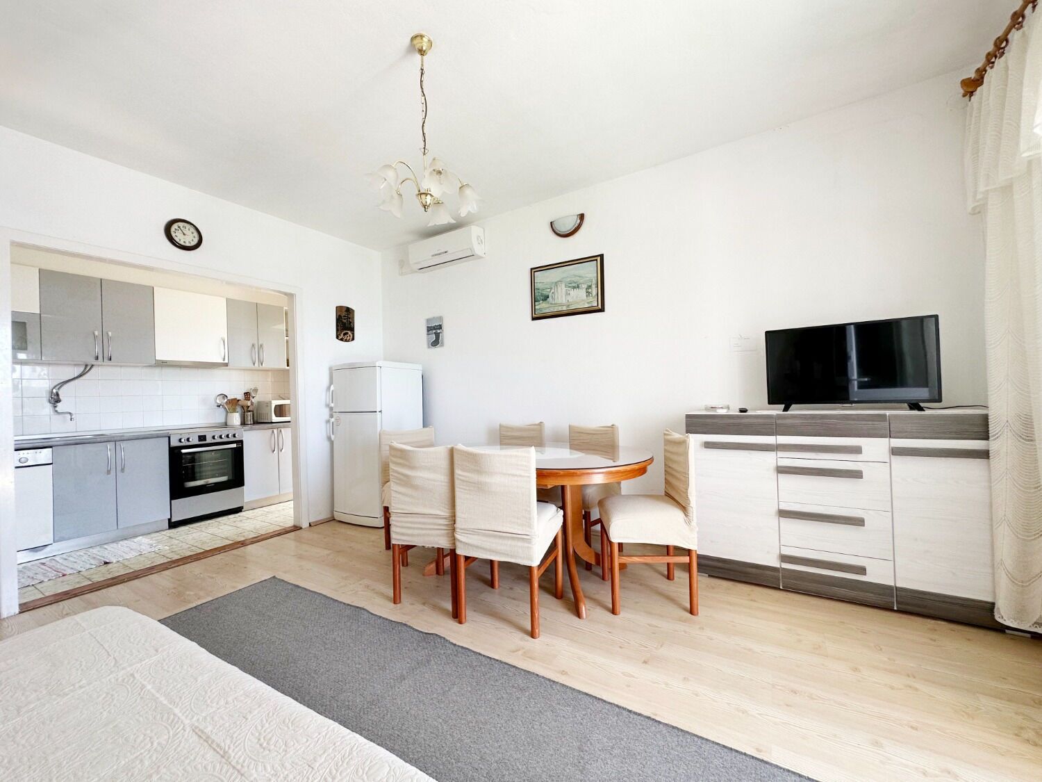 A modern kitchen with white cabinets, a built-in oven, and a refrigerator. There is a dining table with chairs and a TV on a stand.