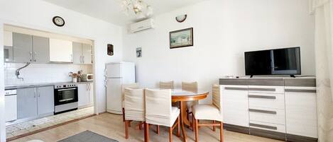 A modern kitchen with white cabinets, a built-in oven, and a refrigerator. There is a dining table with chairs and a TV on a stand.