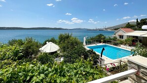 Une piscine avec vue sur la mer, entourée de verdure et bordée d’un bâtiment au toit rouge.