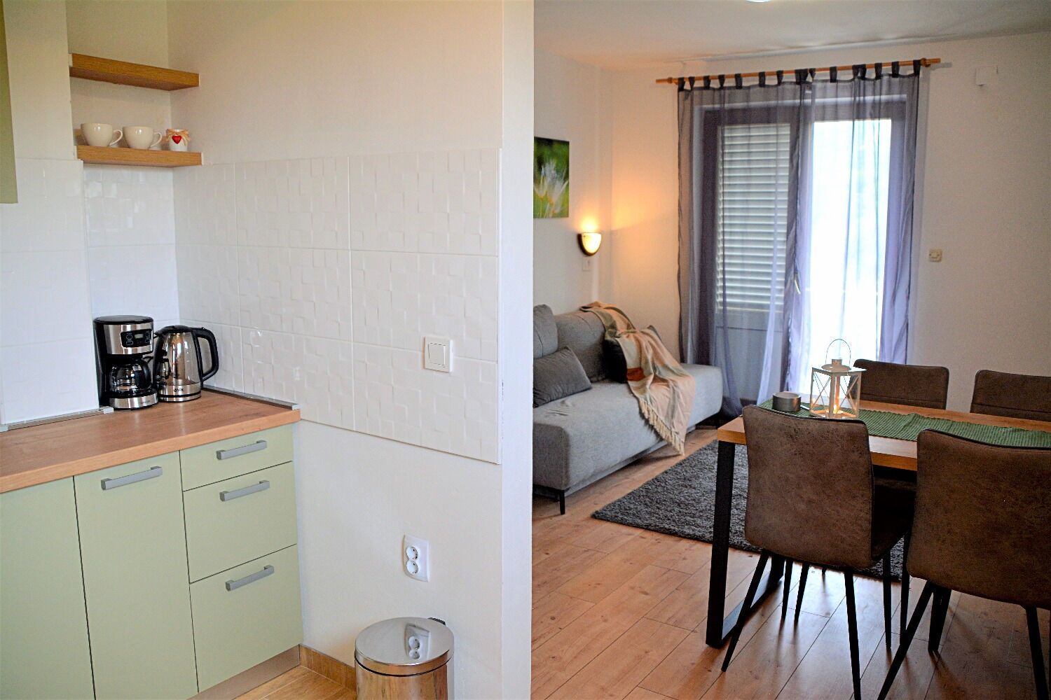 A kitchenette with a coffee maker and a trash bin, a dining area with a table and chairs, and a living area with a sofa and a lamp.