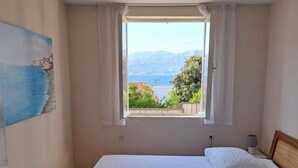 Ein Schlafzimmer mit Bett, einem Fenster mit Vorhängen, einem Bild an der Wand und Blick auf das Meer und die Berge.