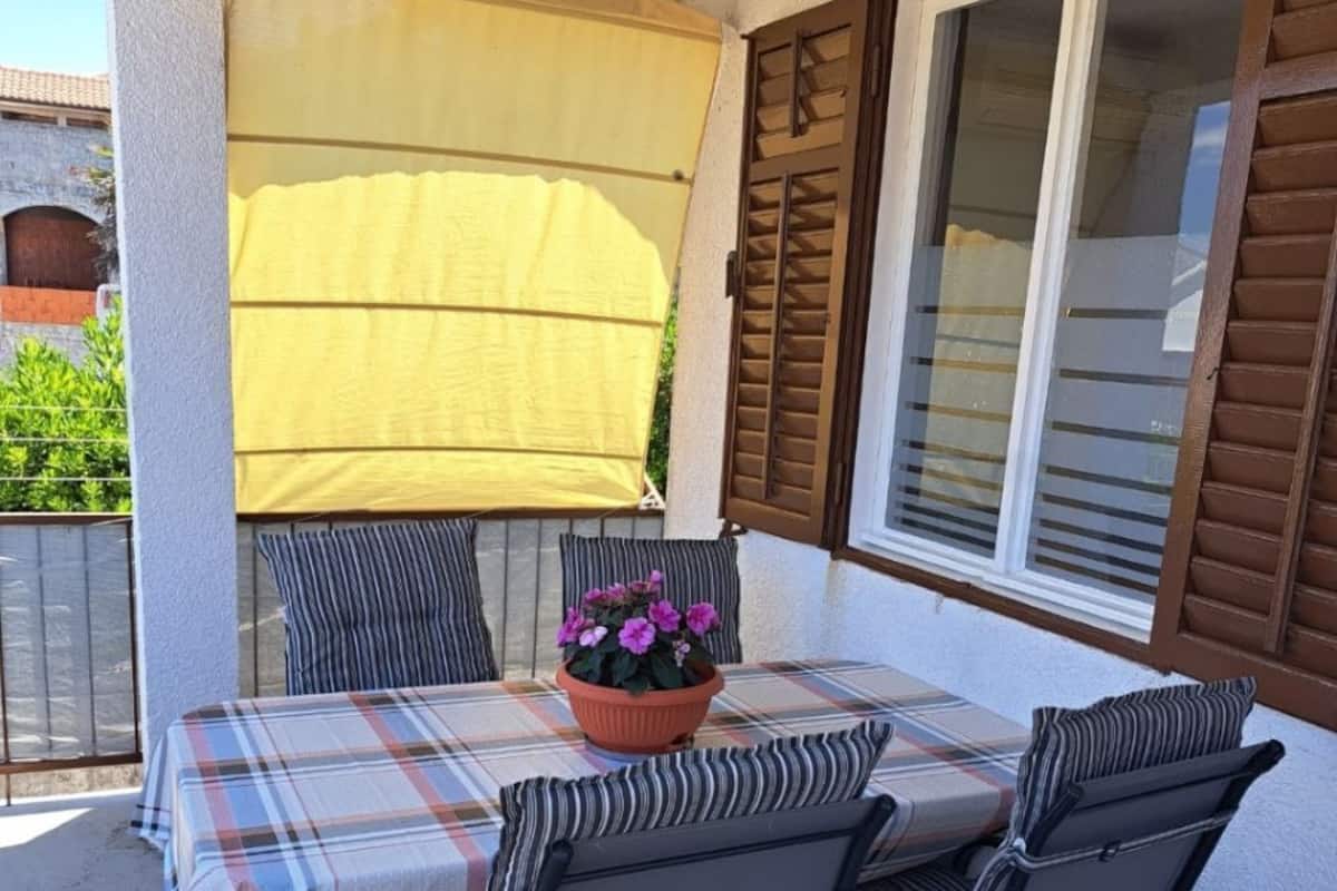 Un patio al aire libre con una mesa preparada para cuatro personas, rodeada de sillas, un toldo amarillo y una planta en maceta con flores moradas.
