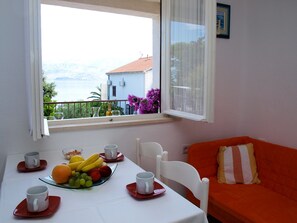 Una habitación con una mesa de comedor preparada para dos personas, una ventana que ofrece vistas a un edificio y montañas, y un sofá naranja con un cojín a rayas.
