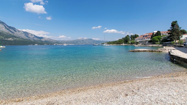 Una spiaggia di ciottoli offre una vista aperta su acque calme, imbarcazioni, una catena montuosa in lontananza e edifici con tetti rossi.