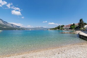 A clear view of a pebble beach leading to calm waters with boats, a distant mountain range, and buildings with red roofs. - Apartment Vedro (Korčula)