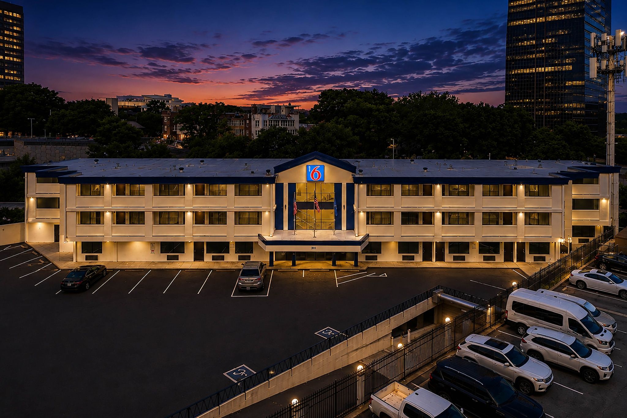 A multi-story hotel with a parking lot, a prominent entrance with a blue sign, and a sunset sky in the background.