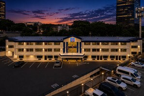 A multi-story hotel with a parking lot, a prominent entrance with a blue sign, and a sunset sky in the background. - 2 Full Beds|Mobil Access|Nsmk|Micfridg (Atlanta)