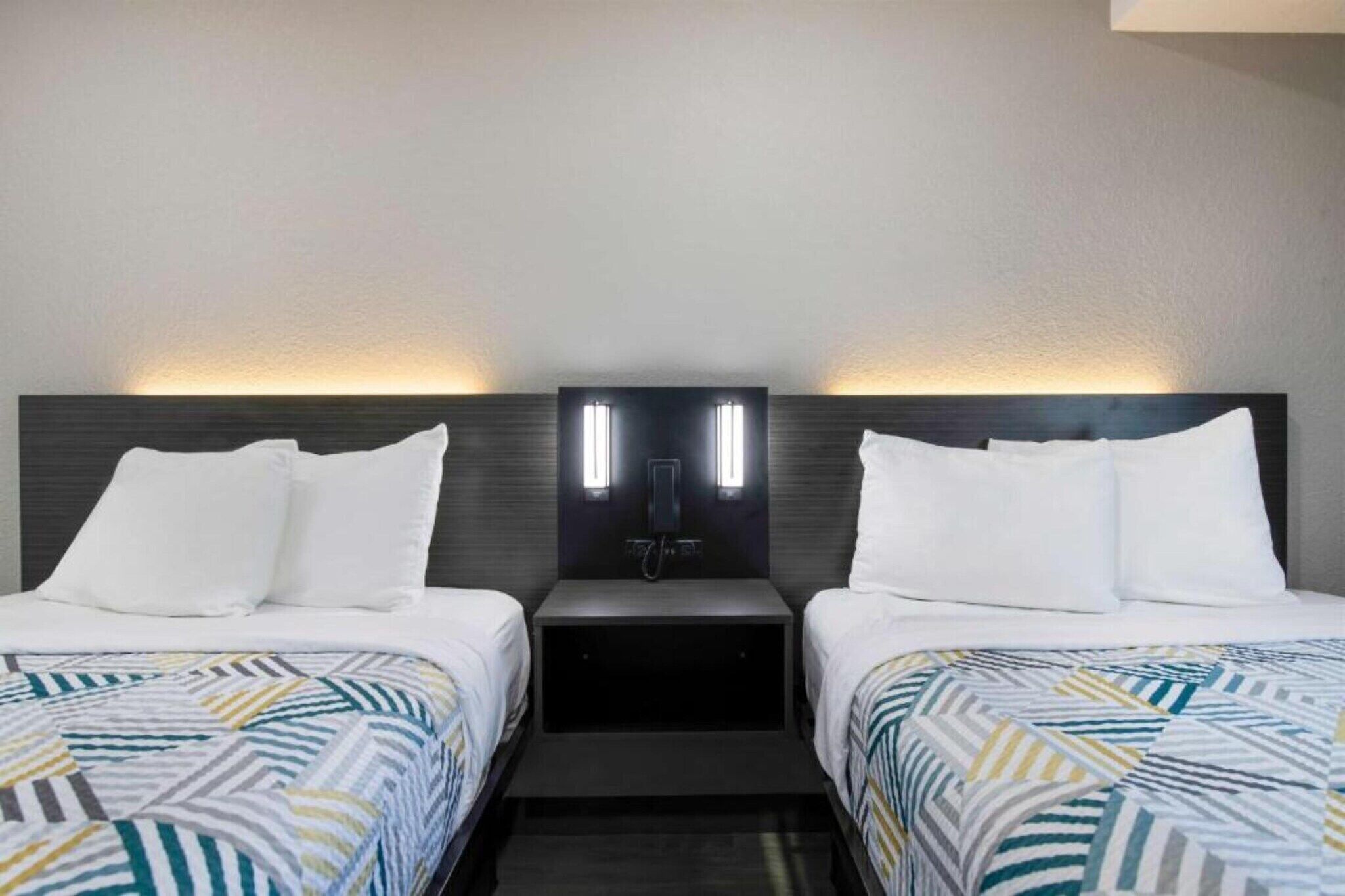 A hotel room with two beds, each with white bedding and a patterned duvet. There are two bedside tables with lamps and a wall-mounted light fixture between the beds.