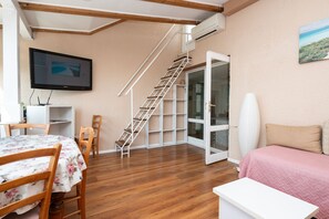 Une pièce avec un escalier en bois, un téléviseur fixé au mur, une table à manger avec des chaises et un lit recouvert d’une couverture rose.