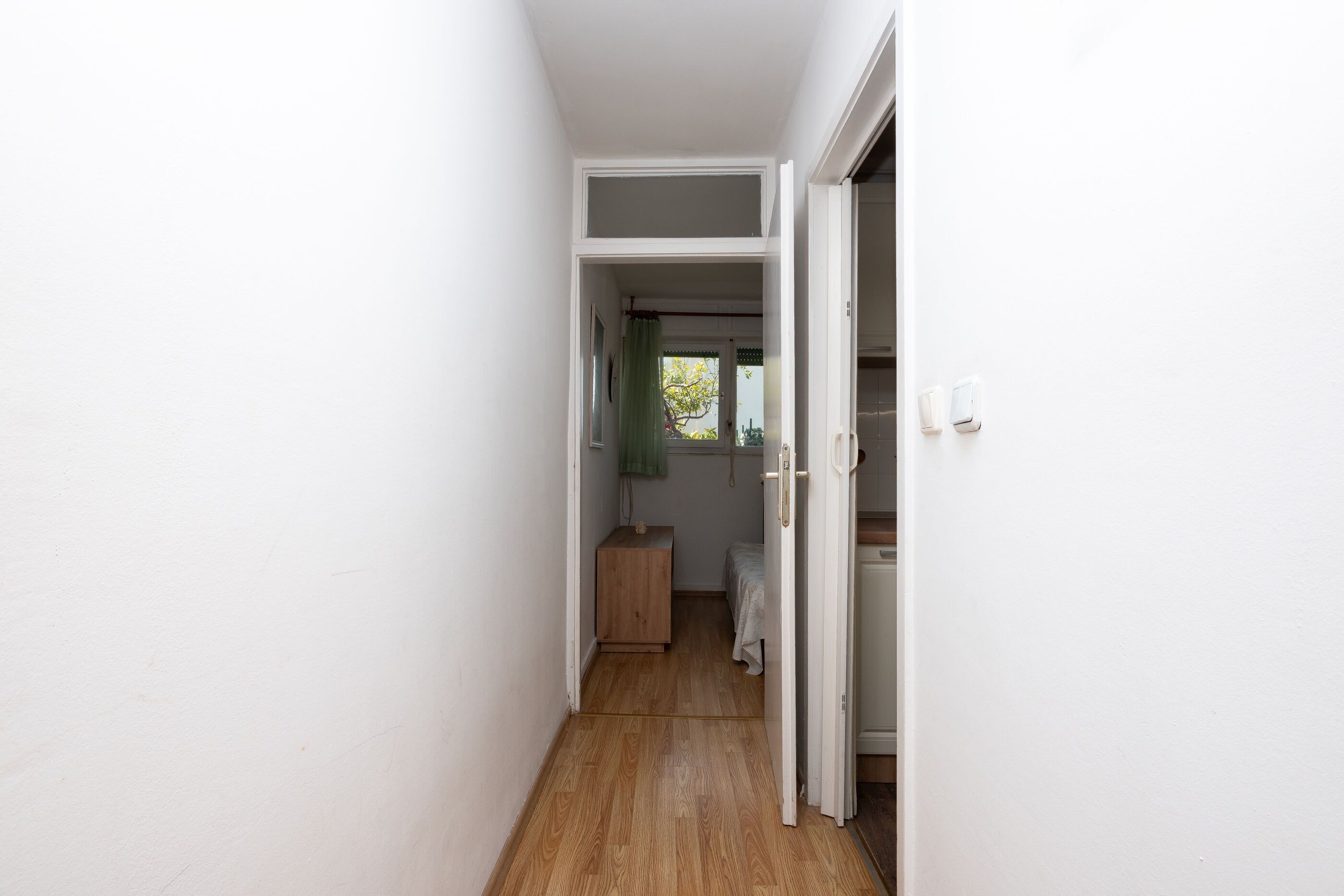 Ein schmaler Korridor mit Holzboden führt zu einem Zimmer mit einem Bett und einem kleinen Schrank.