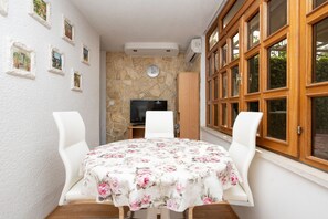Un coin repas avec une table ronde recouverte d’une nappe à motifs floraux, deux chaises blanches, un mur en pierre, une horloge et un téléviseur posé sur un meuble en bois.