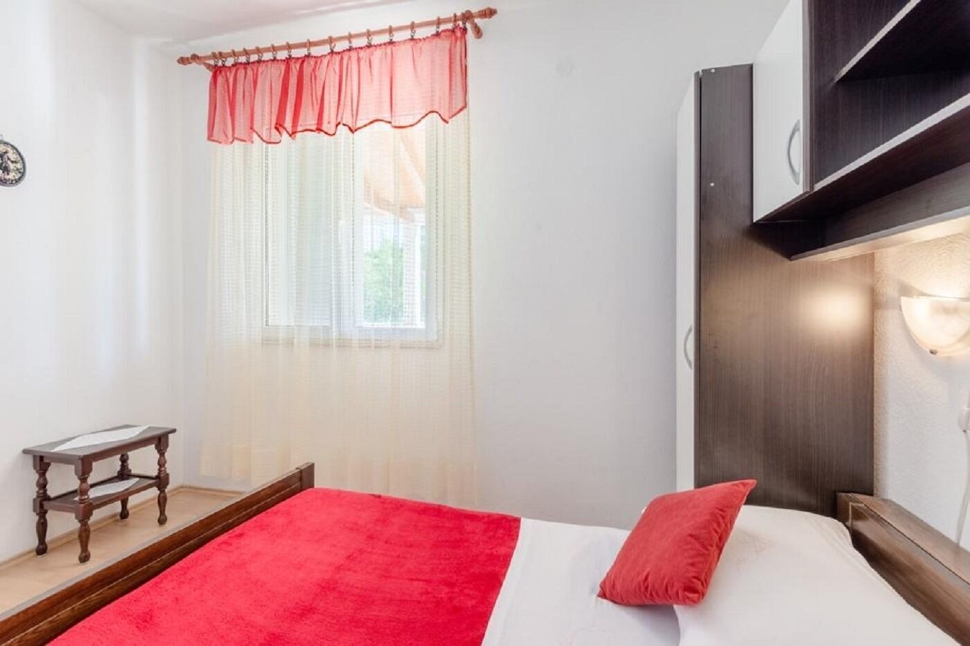 Ein Schlafzimmer mit Bett, Nachttisch, einem Fenster mit durchsichtigen Vorhängen und einem an der Wand montierten Fernseher.
