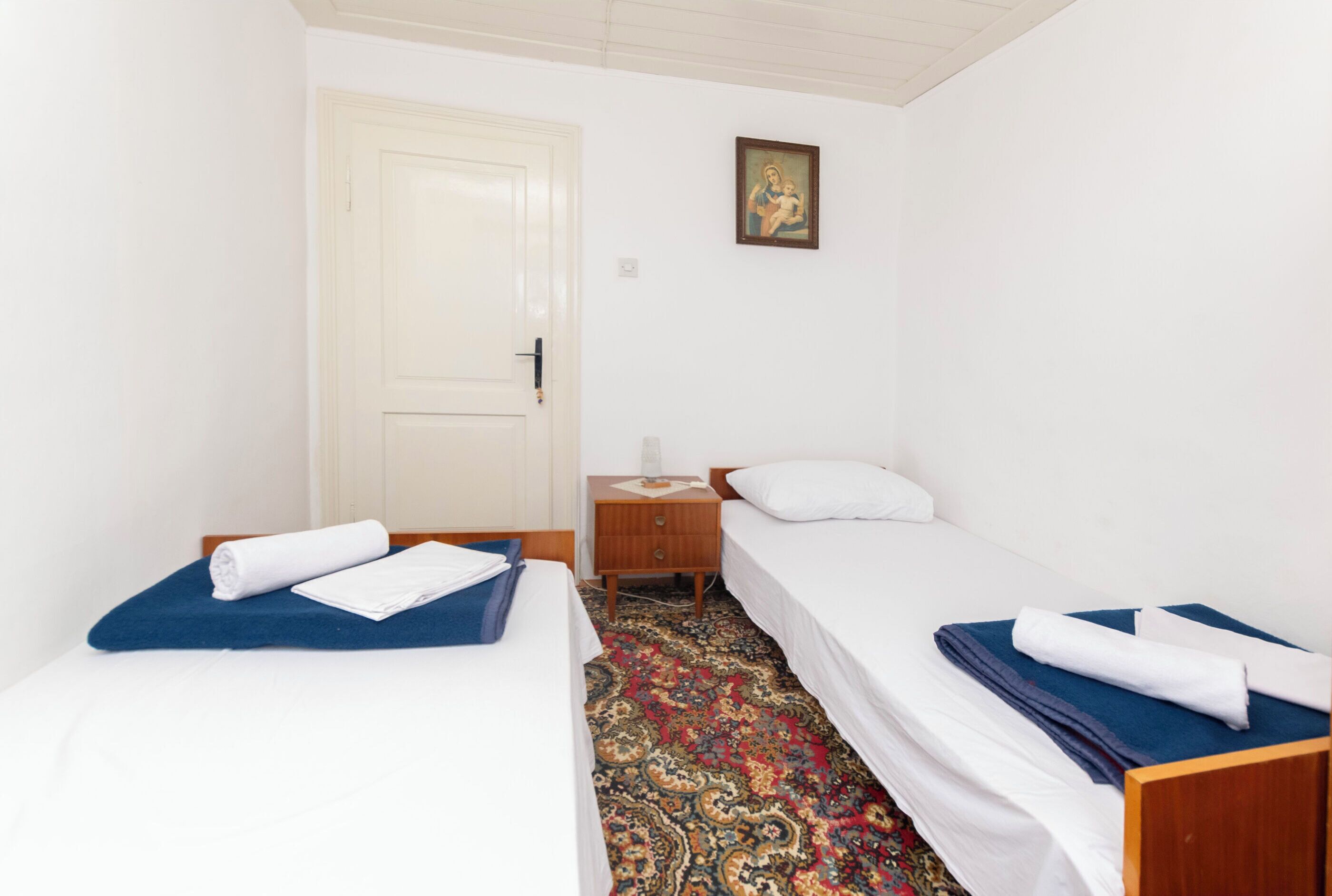 Una habitación pequeña y limpia con dos camas individuales, una mesita de noche de madera, una puerta blanca y un cuadro enmarcado en la pared.