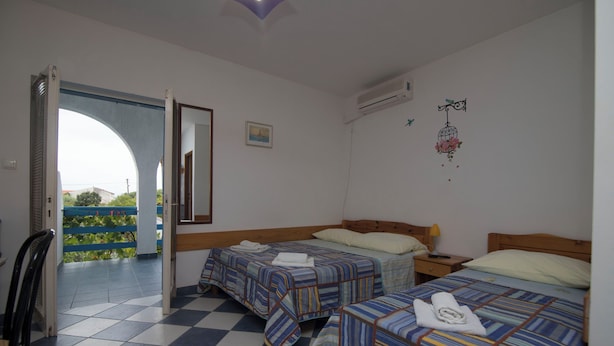 Una camera d'albergo con due letti, pavimento a scacchi, vista attraverso una porta aperta e una parete decorata con una gabbia per uccelli.