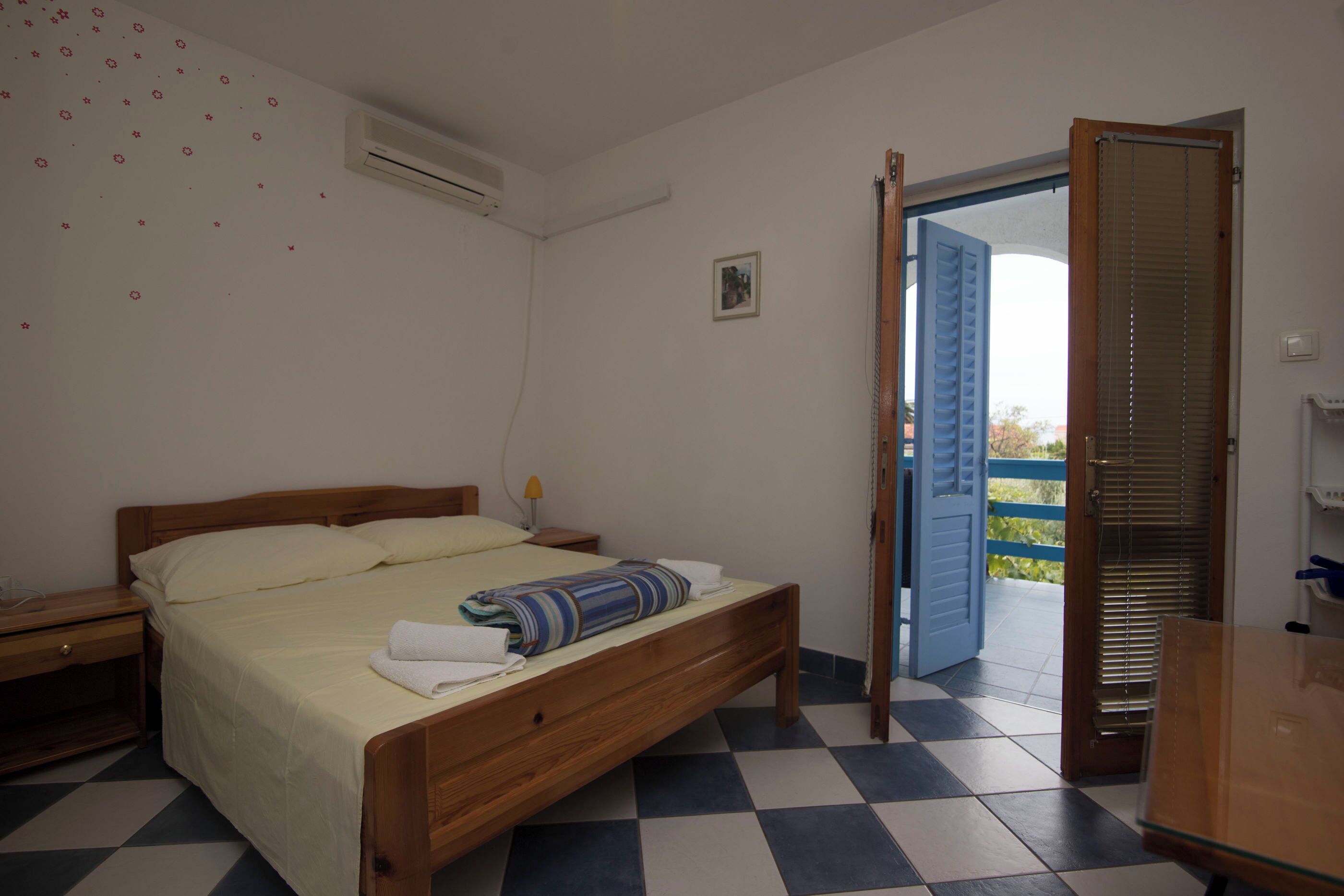 Ein Schlafzimmer mit einem hölzernen Bett, einem Nachttisch, einer Klimaanlage, einem Schachbrettmuster-Boden und einer offenen Tür, die nach draußen führt.