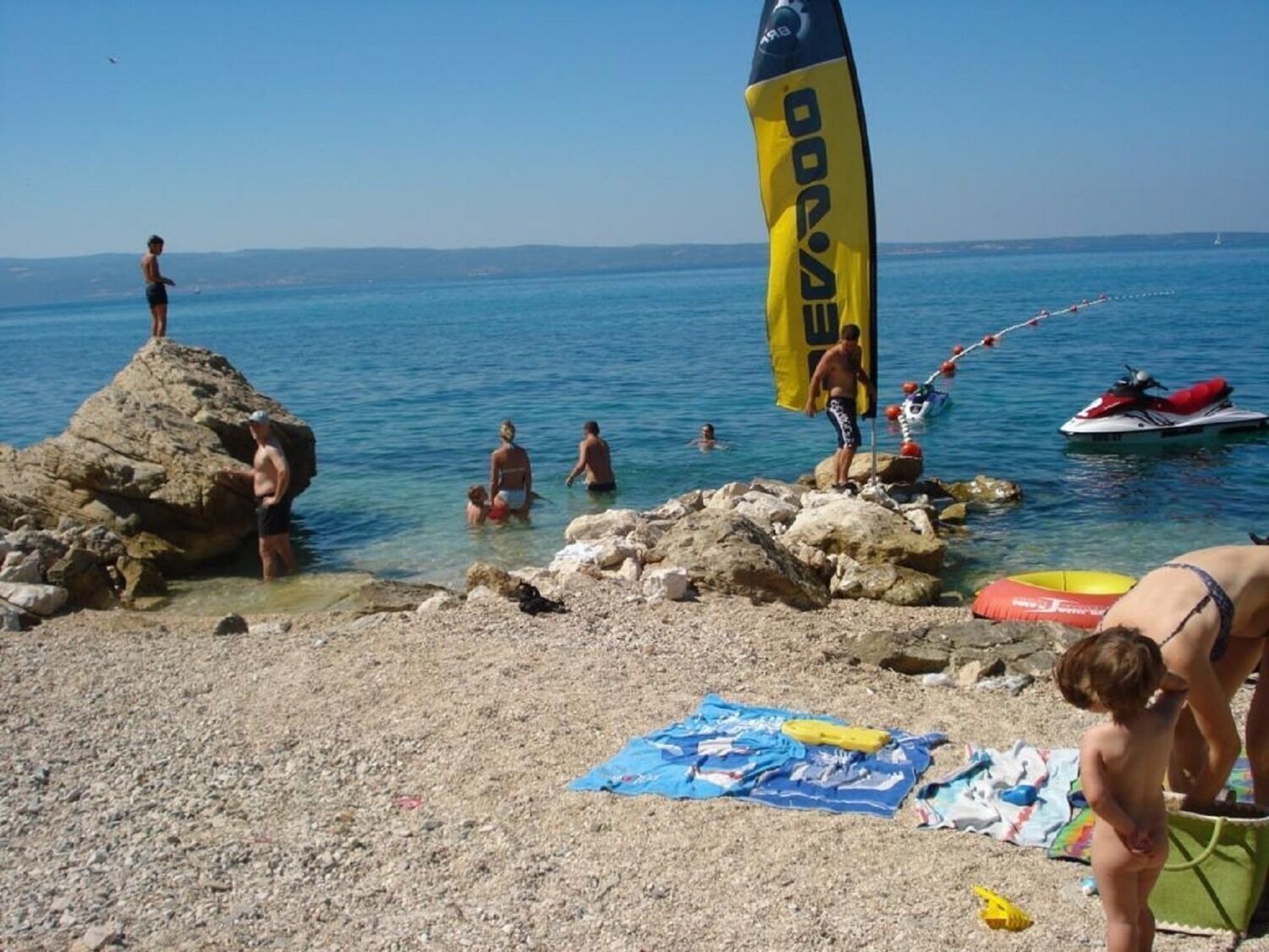 A beach with people swimming and standing on rocks, a jet ski, and a flag.