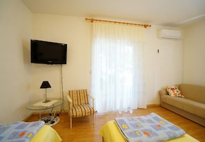 A room with a television, a chair, a sofa, a bed with a floral bedspread, and a window with curtains. - Apartment Marija (Podstrana)