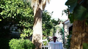 Een betegelde terras met een tafel en stoelen, omringd door weelderige begroeiing en een stenen gebouw op de achtergrond.