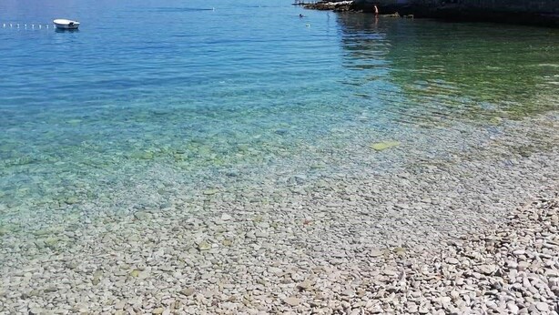 Ett kustområde med klart vatten, en strand med småsten och stenbyggnader med röda tak.