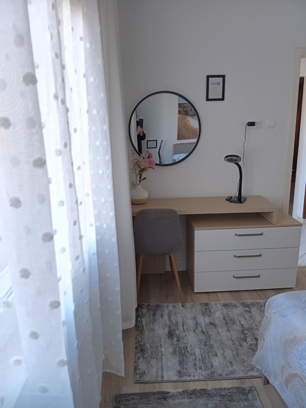 Une chambre à coucher comprenant un lit, un bureau avec des tiroirs, une chaise, un miroir rond, une lampe et une fenêtre avec des rideaux.
