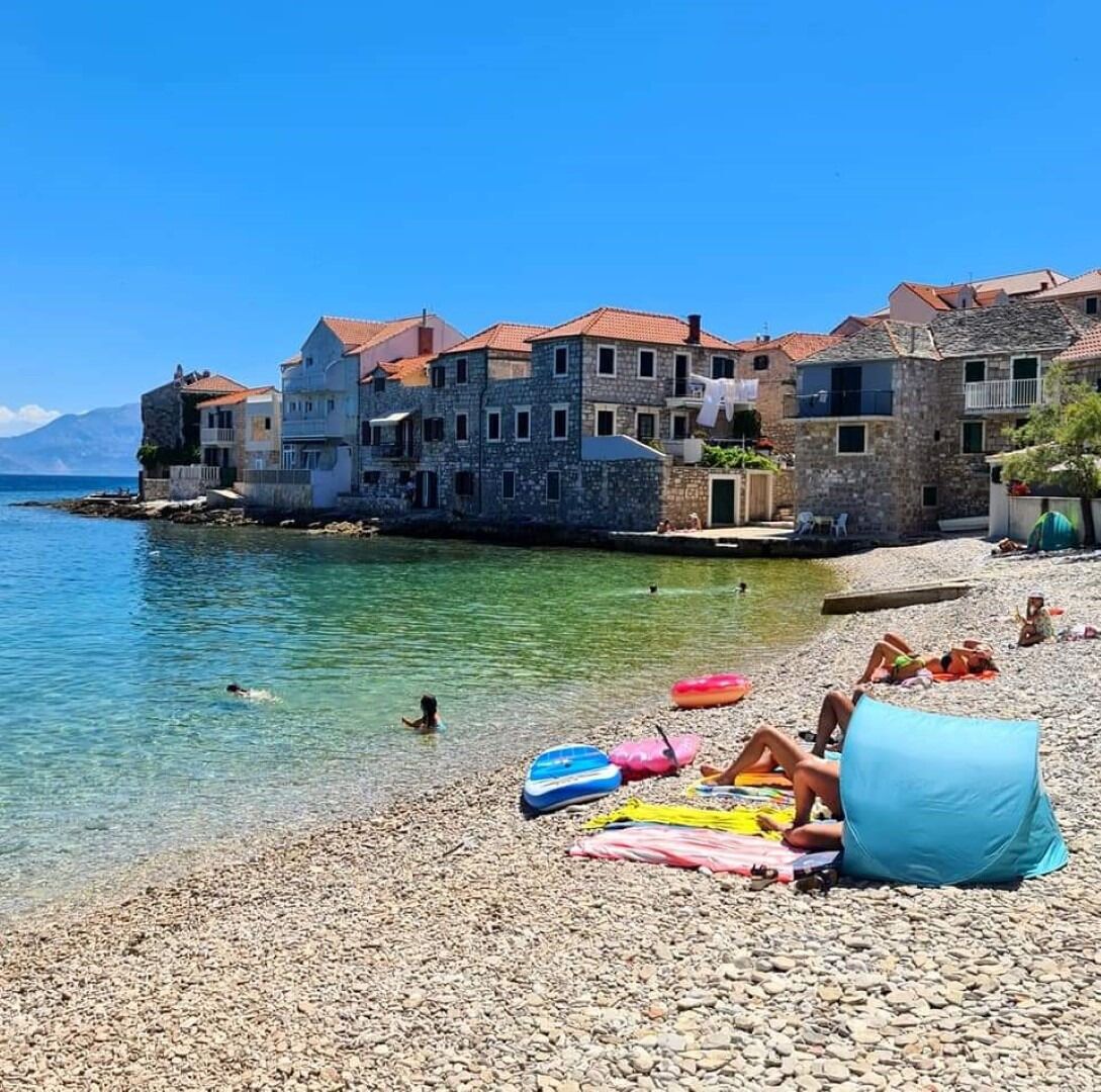 A coastal building with stone walls and red-tiled roofs, situated by a pebbled beach with clear water and a few people swimming.
