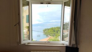 A bedroom with a bed, a window with a view of a body of water and a bridge, and a radiator. - Apartments Kat (Supetar)