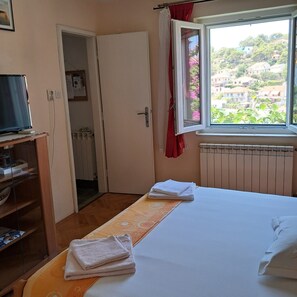 A bedroom with a bed, a window with a view of houses, a radiator, and a TV on a stand. - Apartments Kat (Supetar)