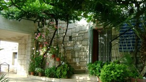 A stone building with a covered entrance, potted plants, and a tiled patio area. - Apartments Kat (Supetar)