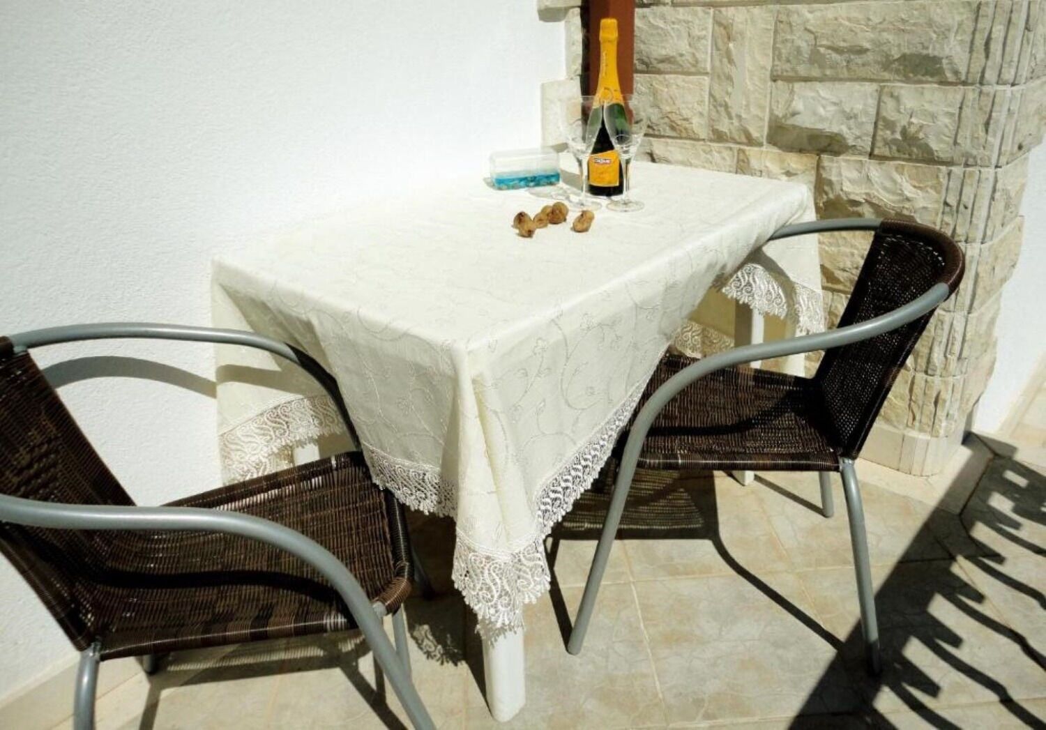A table with a white lace tablecloth, two wicker chairs, a bottle of champagne, and a glass.