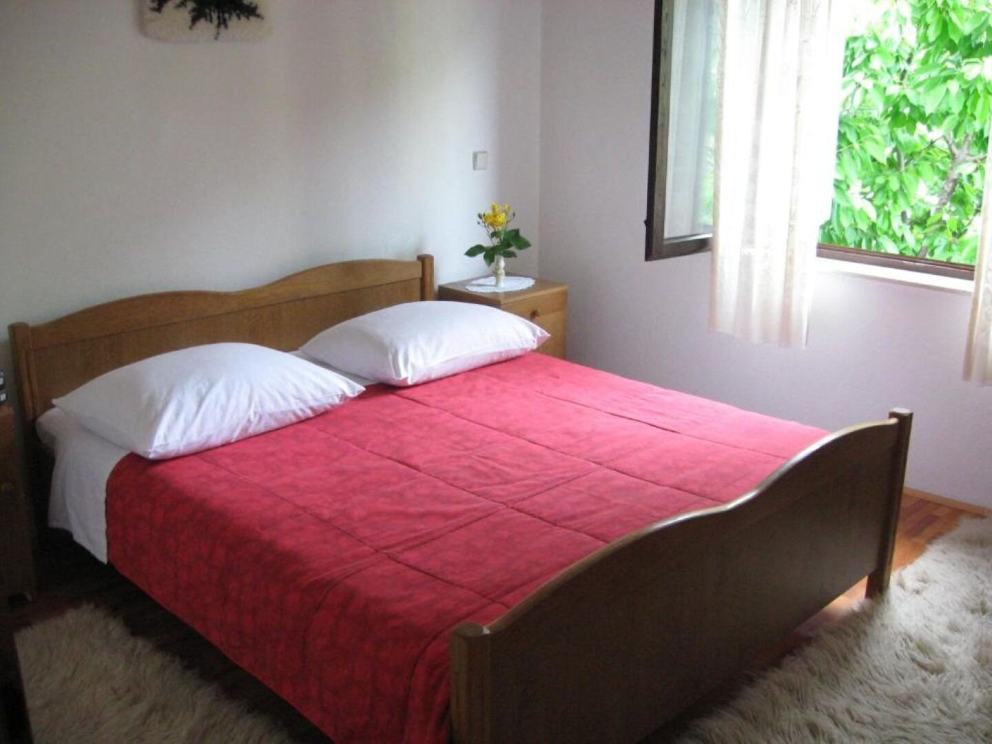 A single bed with a wooden headboard, a red bedspread, and white pillows. There is a nightstand with a small plant and a window with a view of greenery.
