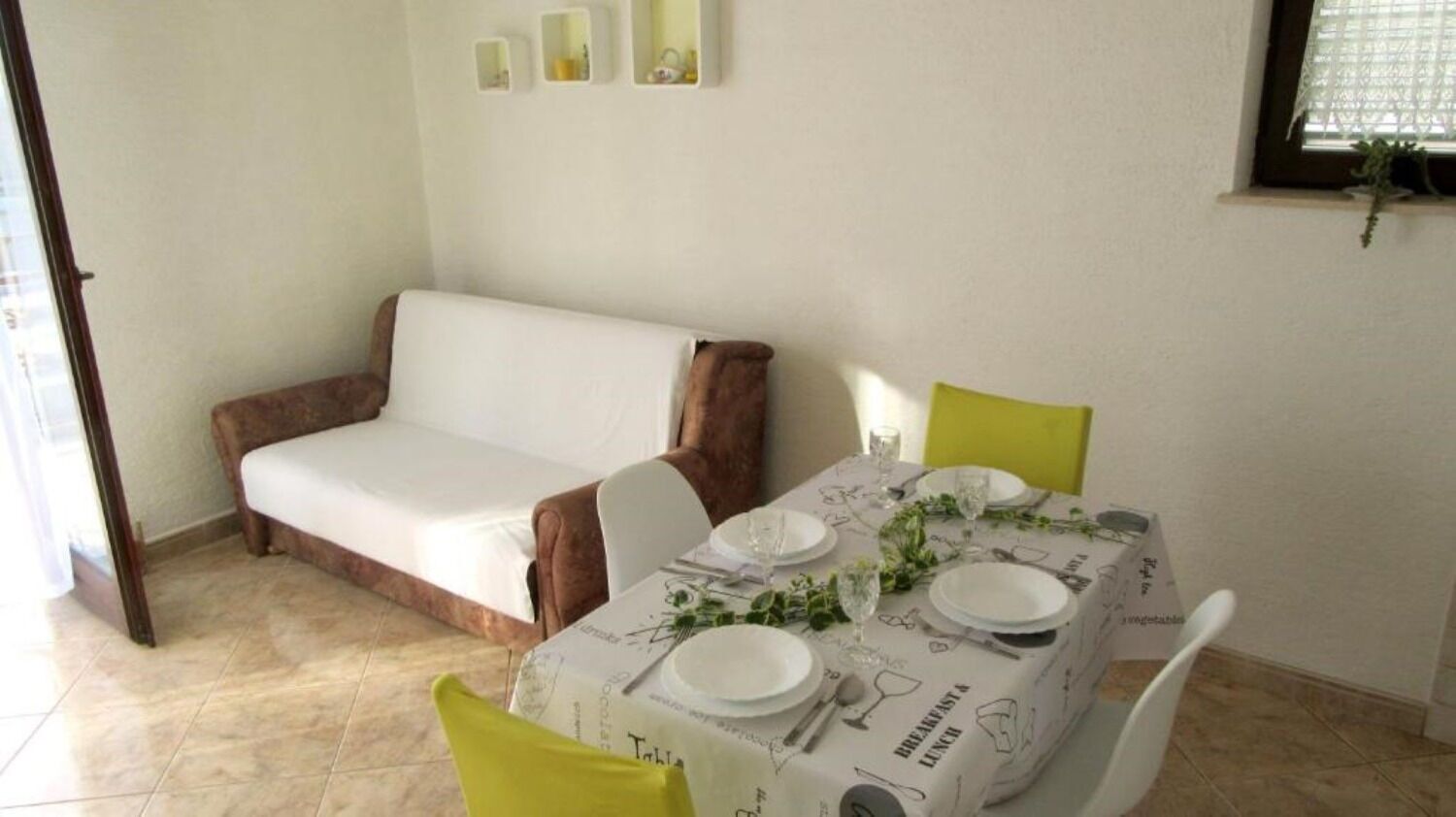 A dining area with a table set for breakfast and lunch, a sofa, and a window with a plant.