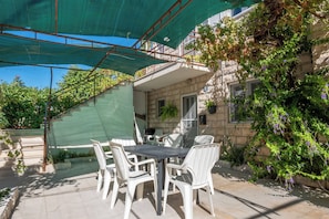 An outdoor patio with a table and chairs, a green canopy, and a stone building with plants. - Apartments Sova (Postira)