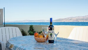 Una mesa con una botella de vino, una copa y un bol de fruta en una terraza con vista al mar.