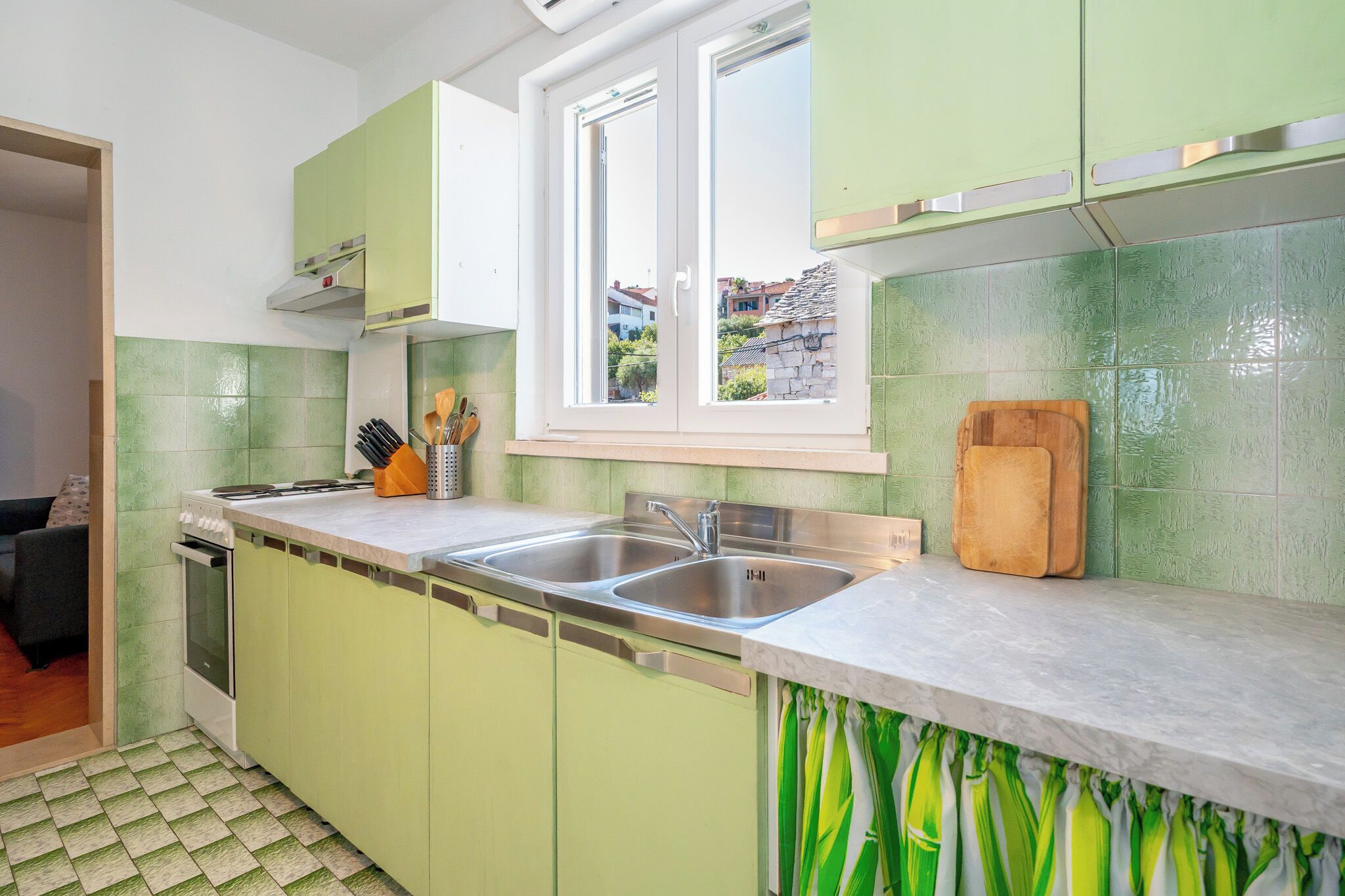 Een keuken met een dubbele spoelbak, groene kastjes en een raam met uitzicht op gebouwen.
