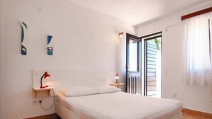 Una camera da letto con un letto, un comodino, una lampada rossa, una parete bianca, una finestra con tende e una porta che dà su una zona esterna.