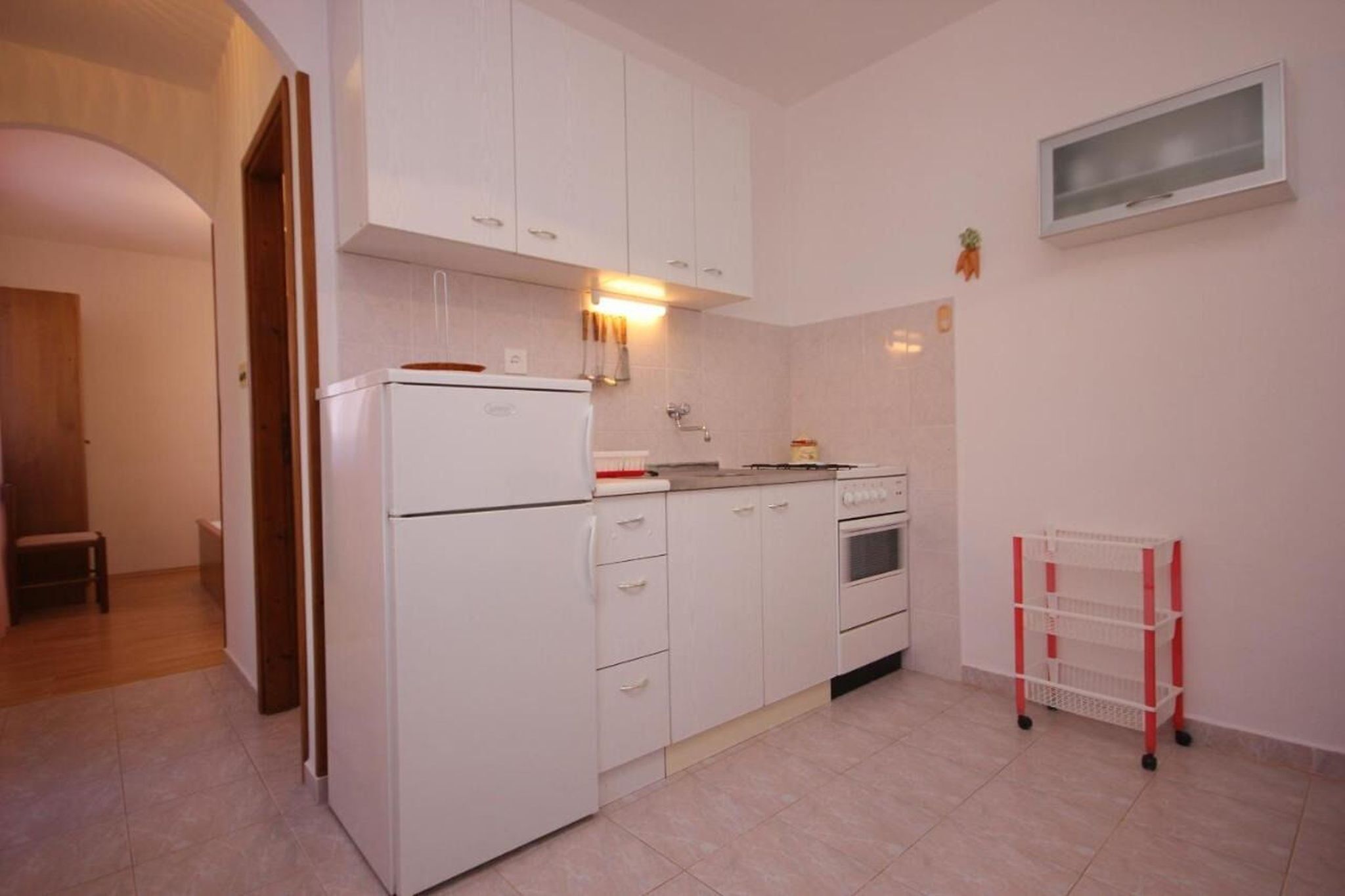 Eine kompakte Küche mit weißen Schränken, einem Kühlschrank, einem Backofen und einem Spülbecken. Über dem Spülbecken befindet sich ein kleines Fenster und ein rot-weiß gestreifter Rollwagen.