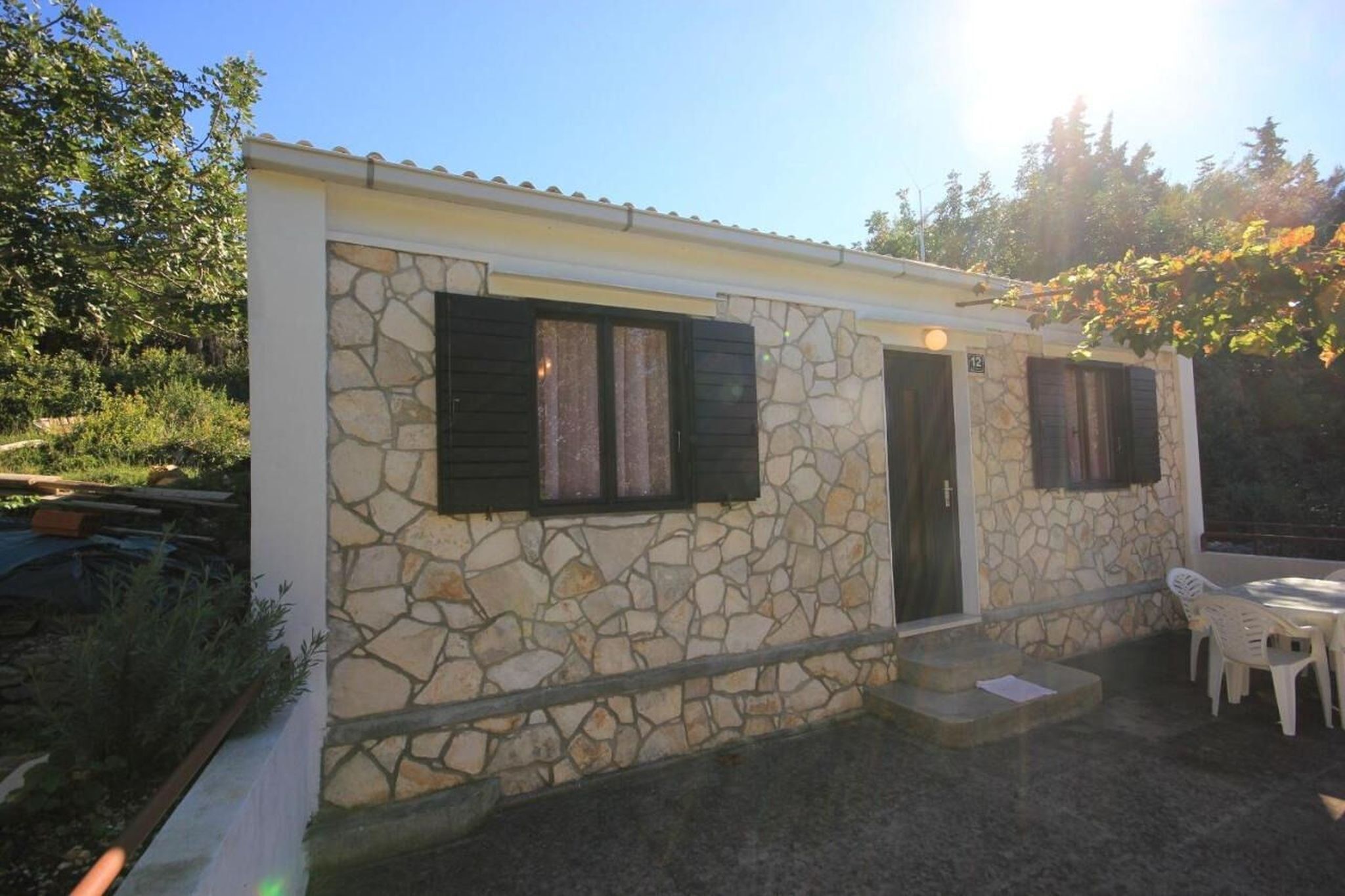 A small stone building with two windows and a door, a patio with a table and chairs, and a number '12' sign on the wall.