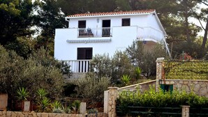 A two-story white building with a tiled roof, surrounded by greenery and a stone wall. - Apartment Ela (Solta)